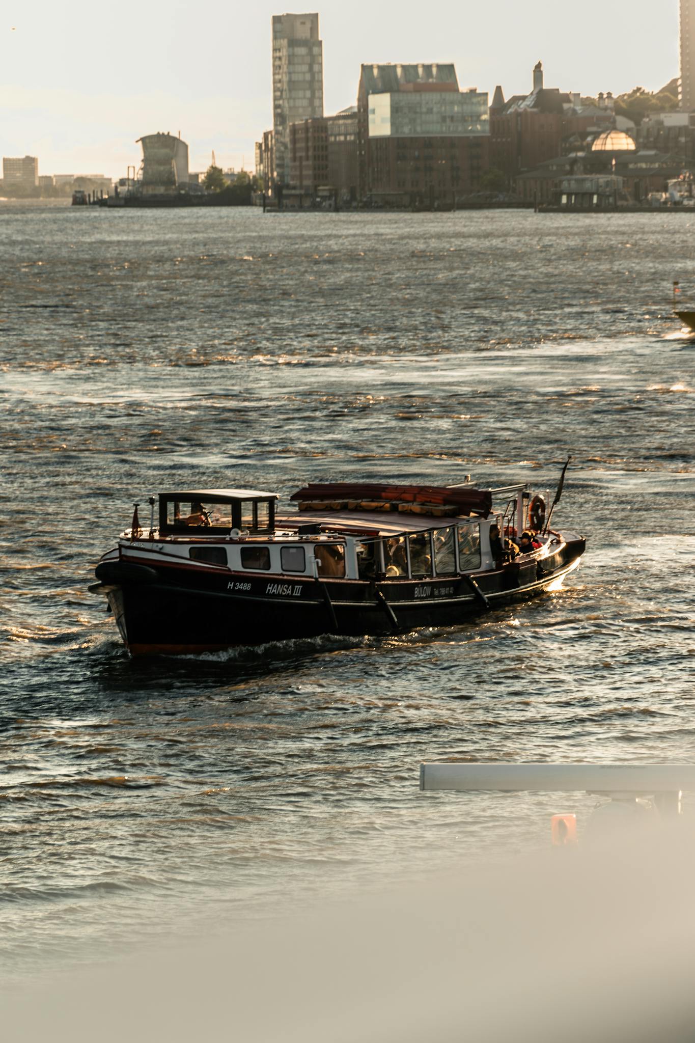 A fishing boat sails through a bustling city harbor at sunset, capturing urban life on the water.