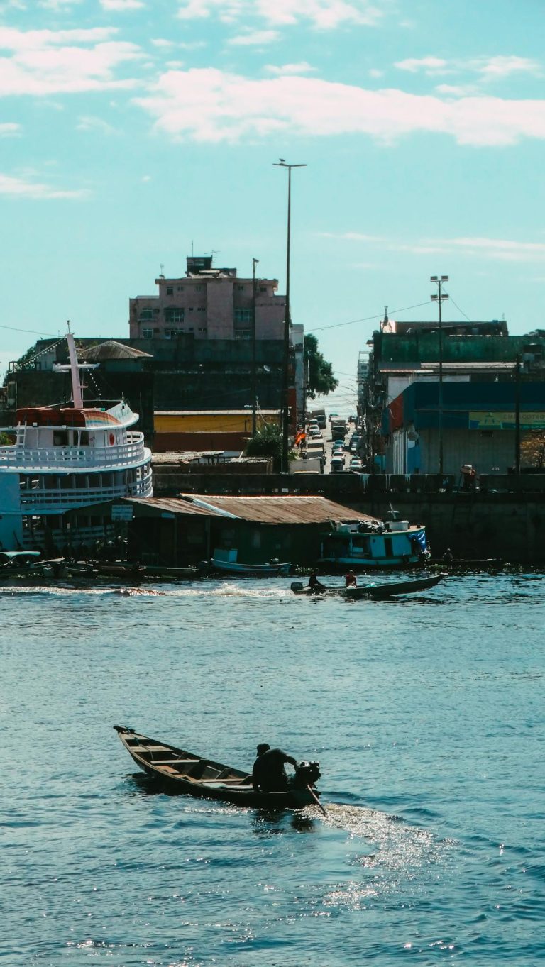 A small motorboat navigates a bustling harbor with urban backdrop under a bright sky.