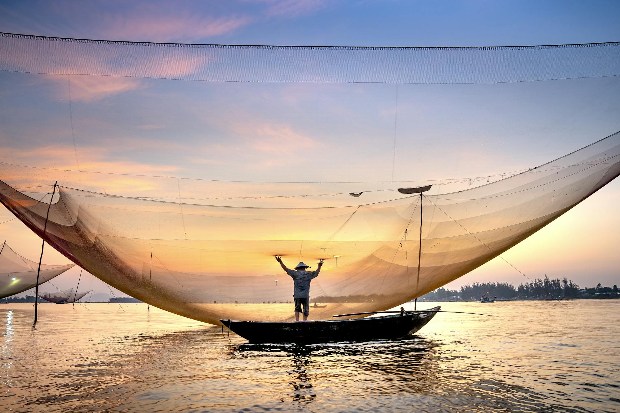 Serene scene of a fisherman casting a large net from a boat at sunset in Vietnam.